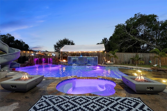 pool at dusk featuring an in ground hot tub, a patio, pool water feature, and a fire pit