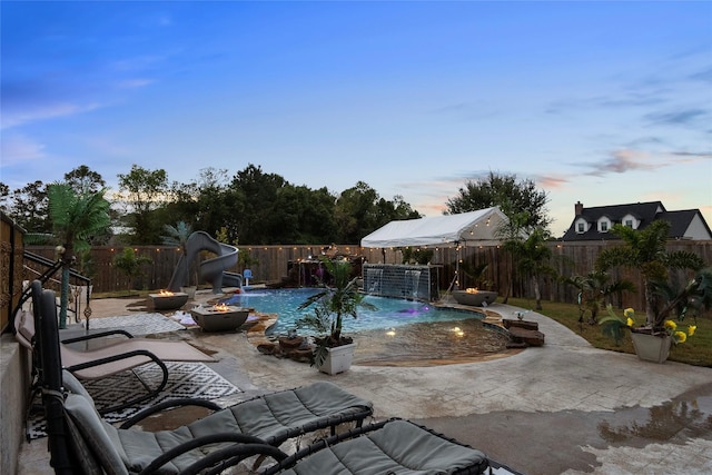 pool at dusk with pool water feature, a water slide, a patio, and an outdoor fire pit