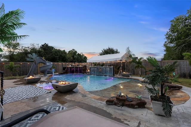 pool at dusk with pool water feature, an outdoor fire pit, a patio area, and a water slide