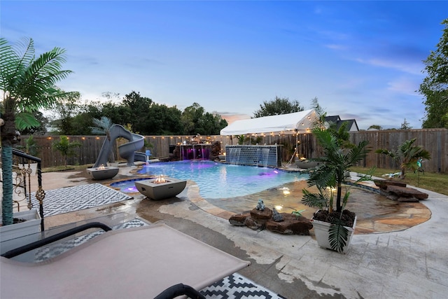 pool at dusk with a patio area, pool water feature, a water slide, and a fire pit