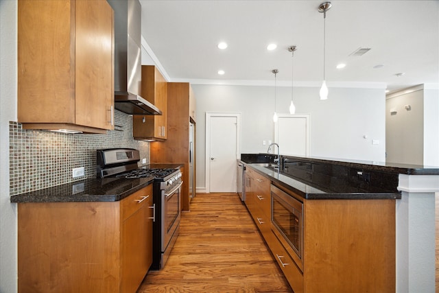 kitchen with dark stone countertops, appliances with stainless steel finishes, kitchen peninsula, wall chimney exhaust hood, and decorative light fixtures