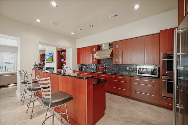 kitchen with a kitchen island with sink, wall chimney exhaust hood, decorative backsplash, a kitchen bar, and stainless steel appliances