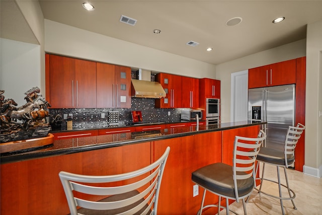 kitchen with backsplash, dark stone counters, a breakfast bar, stainless steel appliances, and wall chimney range hood