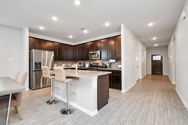 kitchen with light stone countertops, appliances with stainless steel finishes, a breakfast bar, a kitchen island with sink, and light hardwood / wood-style flooring