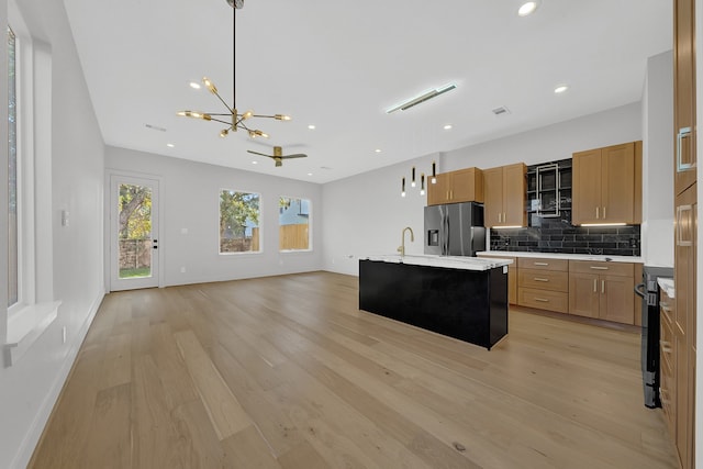 kitchen featuring backsplash, range with electric stovetop, stainless steel fridge, a kitchen island with sink, and light wood-type flooring