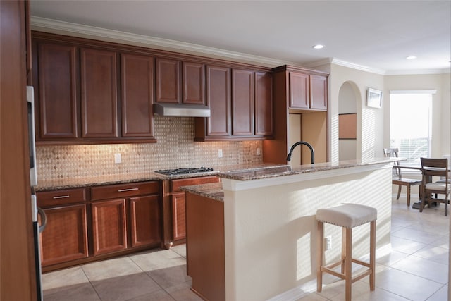 kitchen with light tile patterned flooring, a center island with sink, stainless steel gas cooktop, and dark stone countertops