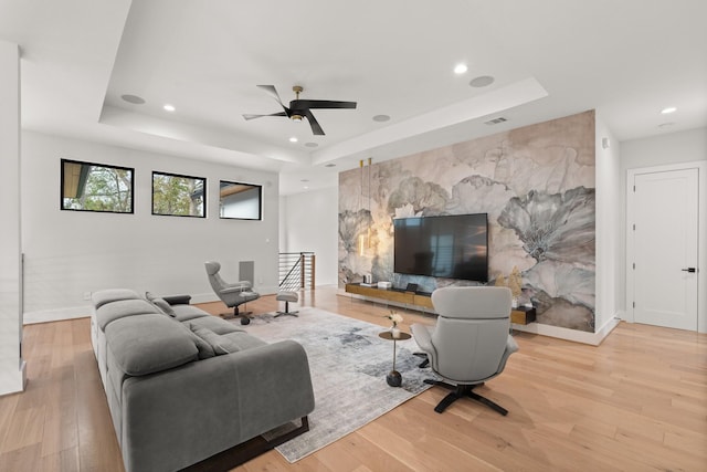 living room with a raised ceiling, ceiling fan, and light wood-type flooring