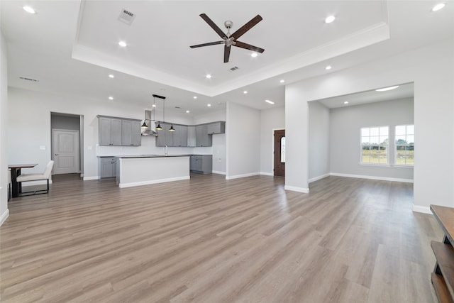 unfurnished living room with sink, ceiling fan, light wood-type flooring, ornamental molding, and a tray ceiling