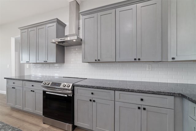 kitchen with wall chimney range hood, light hardwood / wood-style flooring, dark stone counters, stainless steel range with electric stovetop, and decorative backsplash