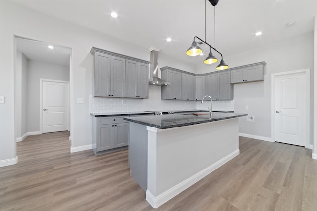 kitchen featuring dark stone countertops, an island with sink, light hardwood / wood-style floors, and wall chimney range hood