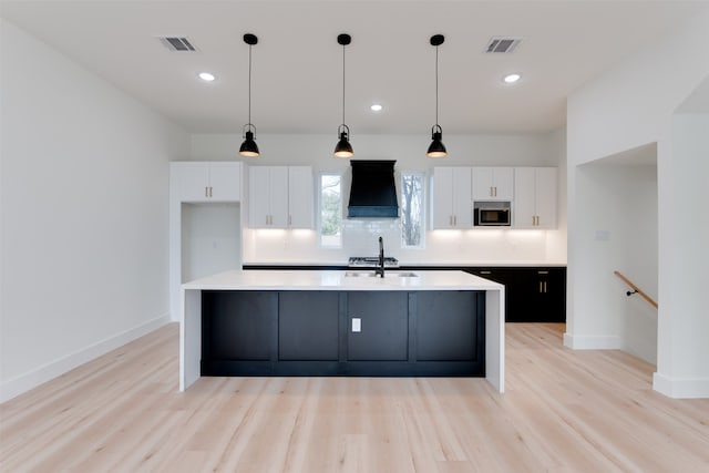 kitchen featuring stainless steel microwave, a kitchen island with sink, white cabinets, light hardwood / wood-style flooring, and custom range hood