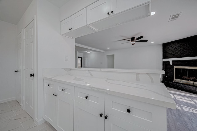 kitchen featuring kitchen peninsula, a brick fireplace, light stone counters, ceiling fan, and white cabinets