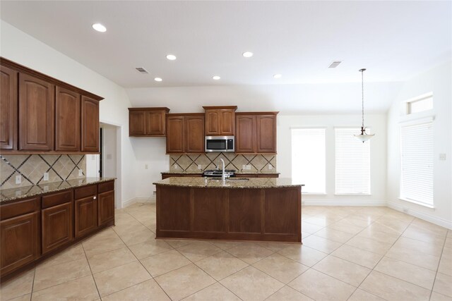 kitchen with tasteful backsplash, light stone countertops, hanging light fixtures, and a kitchen island with sink