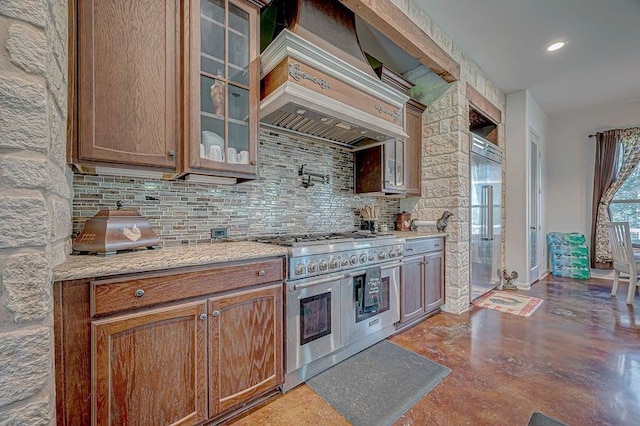 kitchen featuring backsplash, premium range hood, light stone counters, range with two ovens, and concrete floors