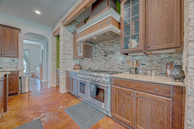 kitchen featuring custom exhaust hood, range with two ovens, light stone counters, and backsplash