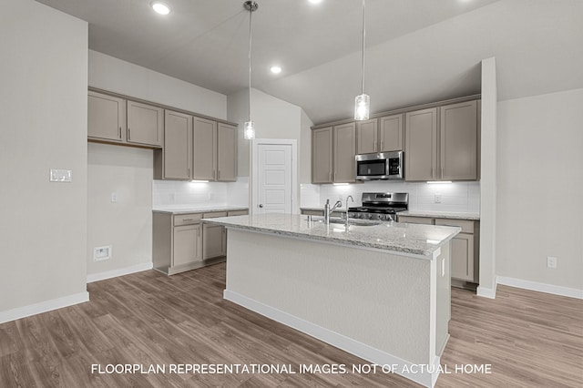 kitchen with hardwood / wood-style floors, a kitchen island with sink, vaulted ceiling, light stone countertops, and appliances with stainless steel finishes