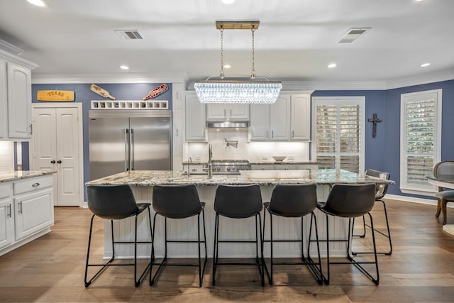 kitchen with stainless steel built in refrigerator, wood-type flooring, white cabinetry, and an island with sink