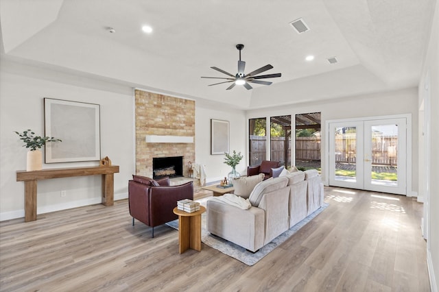 living room featuring french doors, a raised ceiling, a brick fireplace, light hardwood / wood-style flooring, and ceiling fan