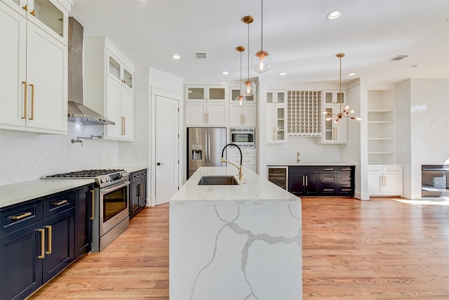 kitchen with wall chimney exhaust hood, pendant lighting, a kitchen island with sink, white cabinets, and appliances with stainless steel finishes