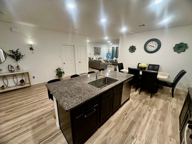 kitchen with dishwasher, a kitchen island with sink, dark stone counters, sink, and light wood-type flooring