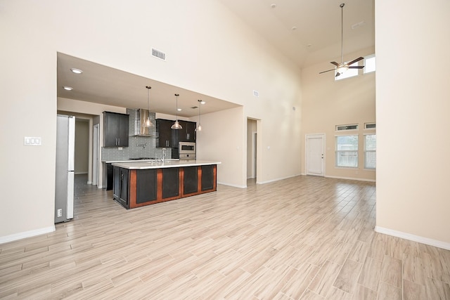 kitchen featuring decorative backsplash, wall chimney exhaust hood, a kitchen island with sink, ceiling fan, and hanging light fixtures