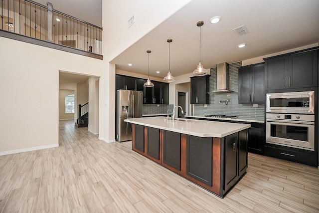 kitchen featuring pendant lighting, a kitchen island with sink, wall chimney range hood, sink, and stainless steel appliances