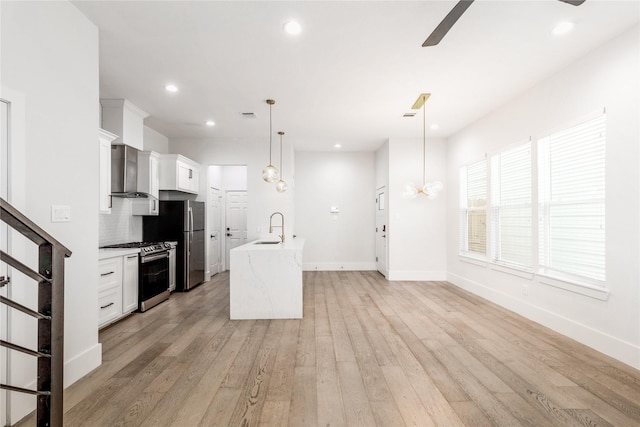 kitchen featuring appliances with stainless steel finishes, wall chimney exhaust hood, decorative light fixtures, white cabinetry, and an island with sink