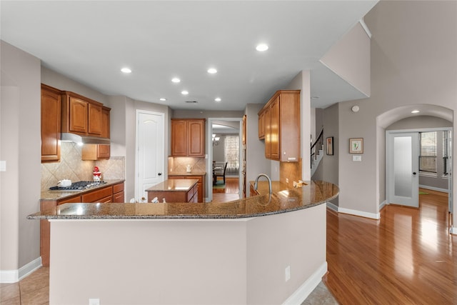 kitchen featuring kitchen peninsula, backsplash, light hardwood / wood-style flooring, dark stone countertops, and stainless steel gas stovetop