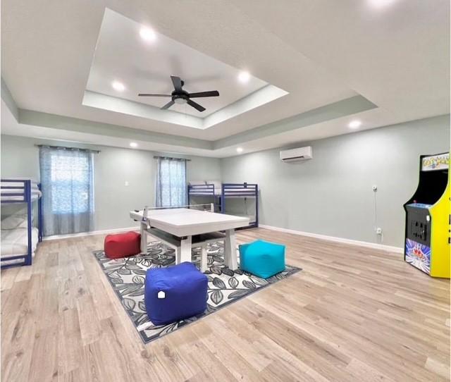 playroom with ceiling fan, light wood-type flooring, a wall unit AC, and a tray ceiling