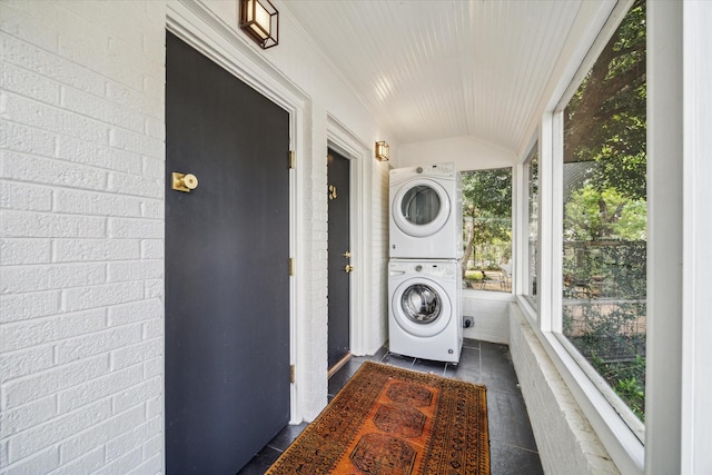 laundry room with stacked washing maching and dryer, dark tile patterned floors, and brick wall