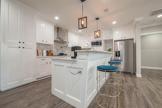 kitchen featuring wall chimney exhaust hood, stainless steel appliances, a kitchen island with sink, pendant lighting, and white cabinets