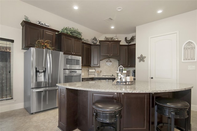 kitchen with decorative backsplash, light stone countertops, dark brown cabinets, stainless steel appliances, and an island with sink