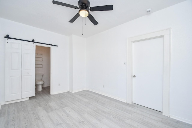 unfurnished bedroom with ceiling fan, a barn door, light wood-type flooring, and connected bathroom