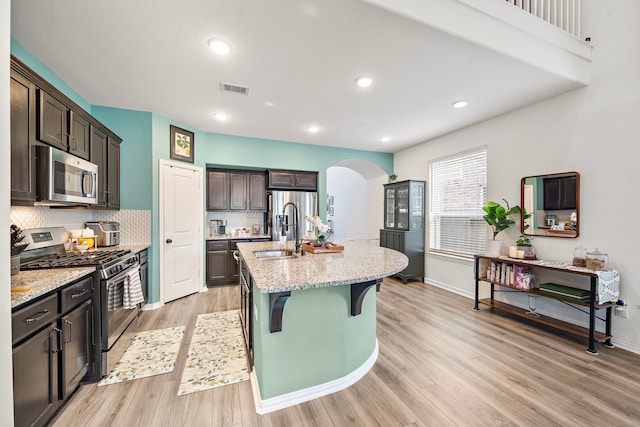 kitchen featuring stainless steel appliances, light stone counters, backsplash, light hardwood / wood-style floors, and a center island with sink