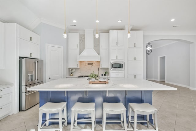 kitchen featuring custom exhaust hood, white cabinetry, a large island with sink, stainless steel appliances, and light stone countertops