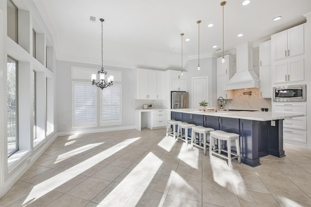 kitchen featuring pendant lighting, stainless steel appliances, custom range hood, white cabinets, and a large island with sink