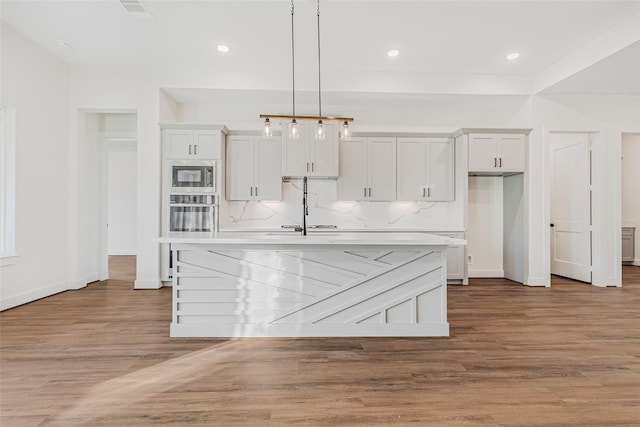 kitchen with pendant lighting, stainless steel microwave, a kitchen island with sink, white cabinets, and light hardwood / wood-style floors