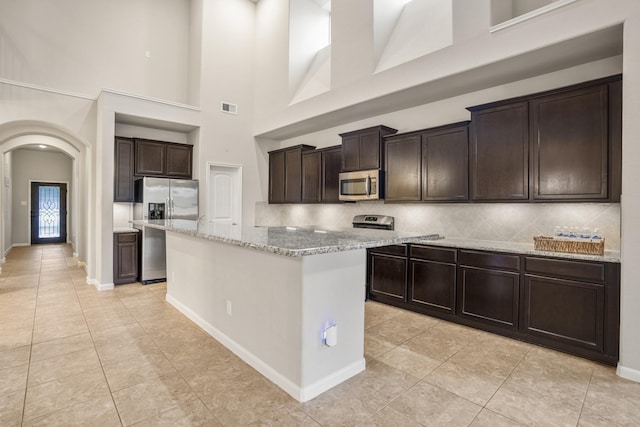 kitchen with light stone countertops, appliances with stainless steel finishes, dark brown cabinetry, a high ceiling, and a center island