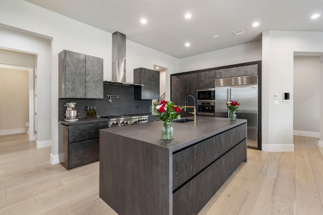 kitchen with sink, wall chimney range hood, light hardwood / wood-style flooring, built in appliances, and a kitchen island with sink