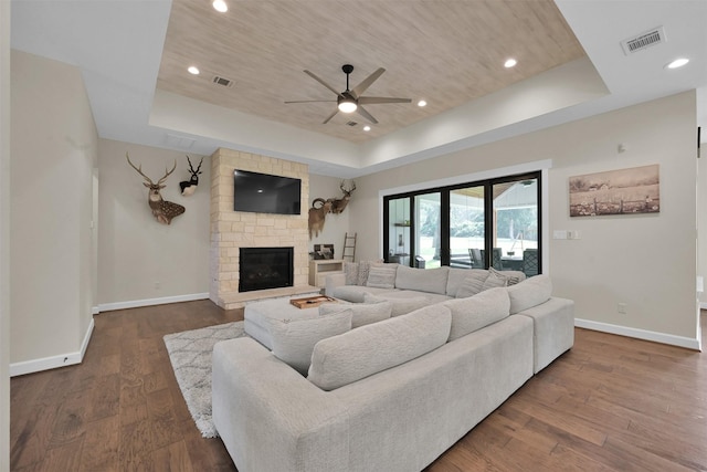 living room featuring a raised ceiling, a stone fireplace, ceiling fan, and dark wood-type flooring