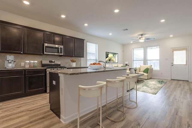 kitchen with stone counters, a center island with sink, light wood-type flooring, dark brown cabinetry, and stainless steel appliances