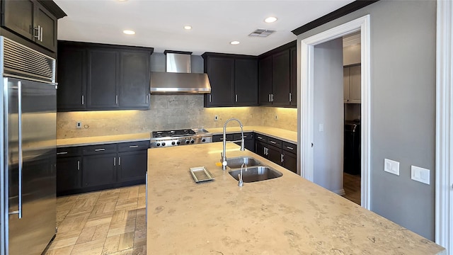 kitchen with decorative backsplash, appliances with stainless steel finishes, sink, wall chimney range hood, and an island with sink