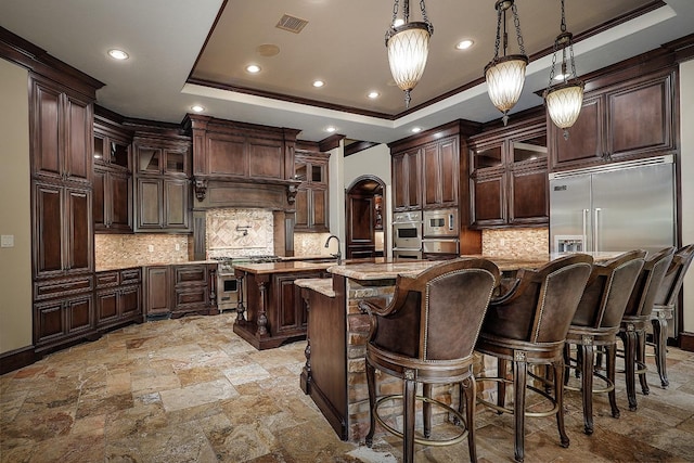 kitchen featuring dark brown cabinets, a tray ceiling, built in appliances, hanging light fixtures, and an island with sink