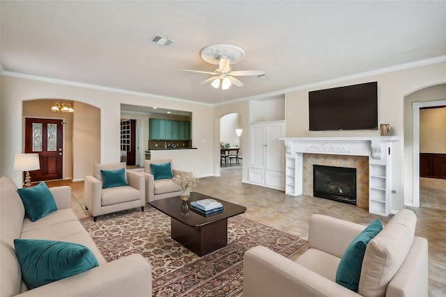 tiled living room with crown molding, ceiling fan with notable chandelier, and a tile fireplace