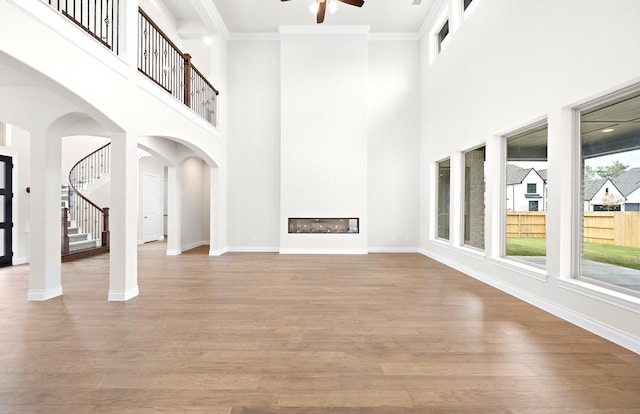 unfurnished living room with ceiling fan, a towering ceiling, crown molding, and light hardwood / wood-style flooring