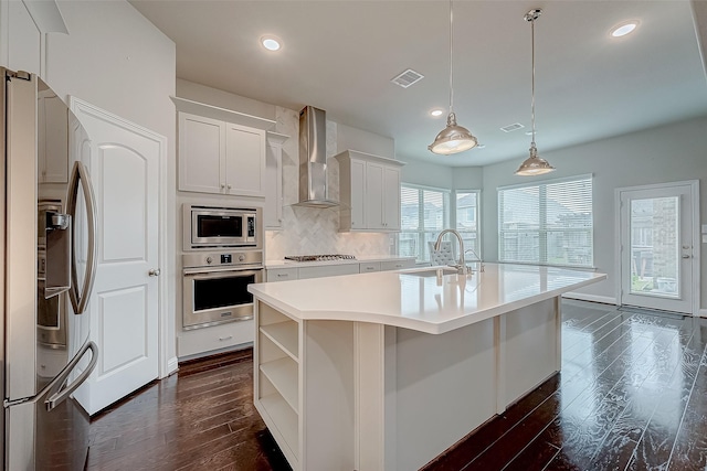 kitchen with a center island with sink, wall chimney range hood, sink, appliances with stainless steel finishes, and white cabinetry