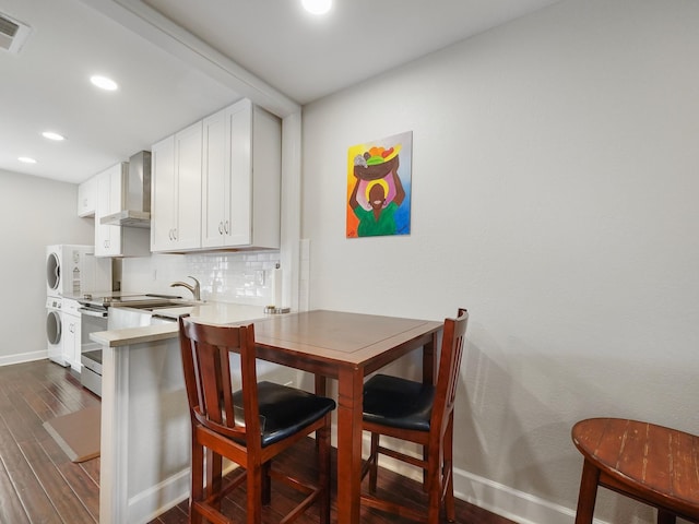 kitchen featuring tasteful backsplash, stacked washing maching and dryer, stainless steel range with electric stovetop, wall chimney range hood, and white cabinetry