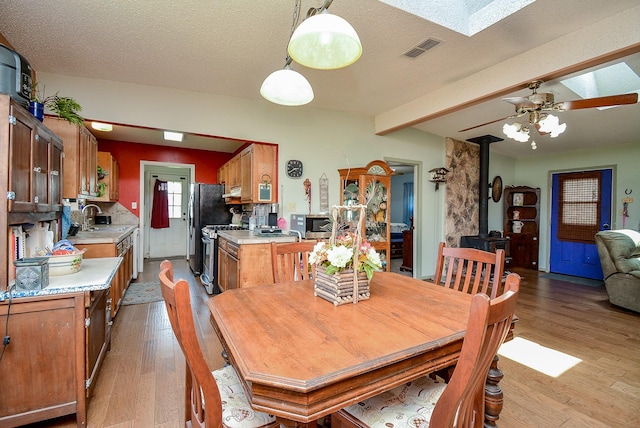 dining room featuring light wood-type flooring, a skylight, a textured ceiling, ceiling fan, and a wood stove