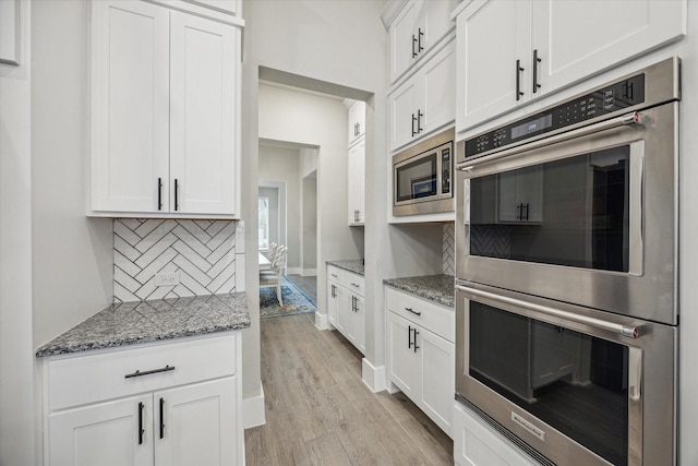 kitchen featuring light wood-type flooring, tasteful backsplash, stainless steel appliances, dark stone countertops, and white cabinets