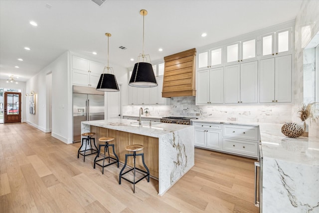 kitchen with appliances with stainless steel finishes, custom range hood, a kitchen island with sink, light hardwood / wood-style flooring, and white cabinets